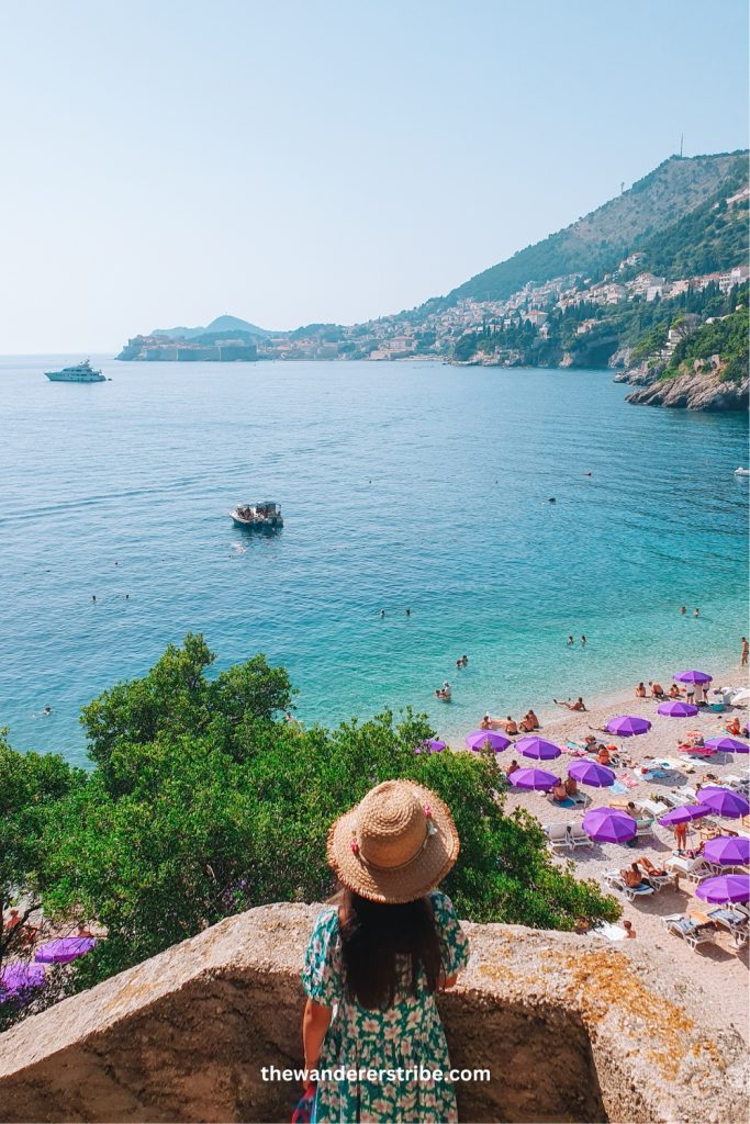 Girl in a straw hat and floral dress looking out over Banje Beach in Dubrovnik, Croatia, with purple umbrellas on the sand and a view of the Adriatic Sea and Old Town in the distance.