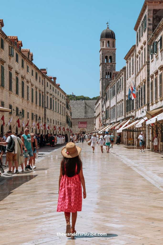Young girl in a pink dress and straw hat walking along the polished stone street of Stradun in Dubrovnik’s Old Town, surrounded by historic buildings and tourists.