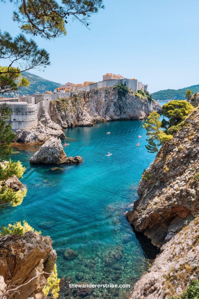View of kayakers paddling through clear turquoise waters beneath the dramatic cliffs and stone walls of Dubrovnik during a Croatia road trip.
