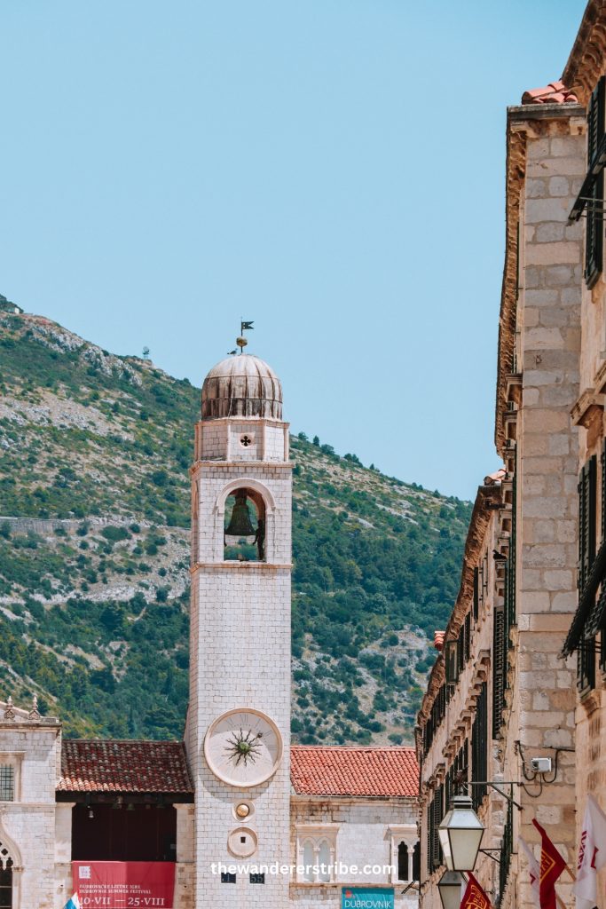 Bell tower with a clock on Stradun street in Dubrovnik Old Town and hills in the background.
