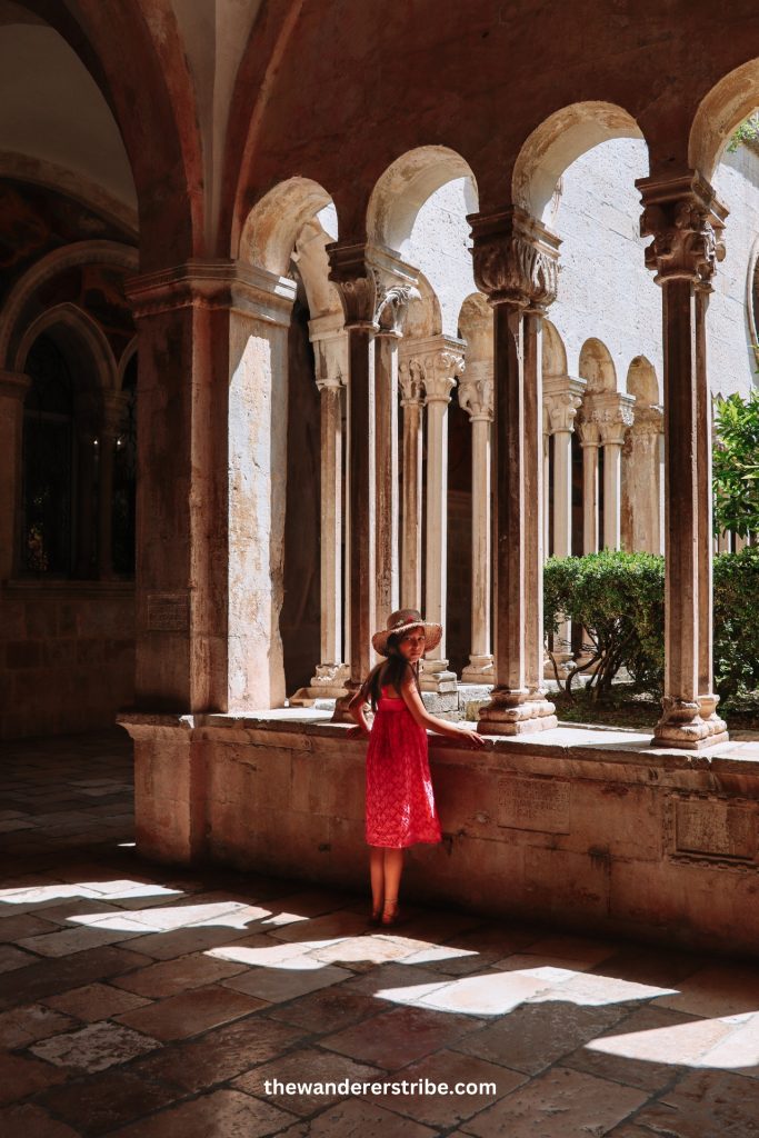 Girl wearing a straw hat and red dress looking at the columns in Dubrovnik’s Franciscan cloister.