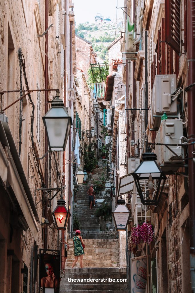 Narrow stepped alley in Dubrovnik Old Town lined with stone buildings, hanging lanterns, and plants.
