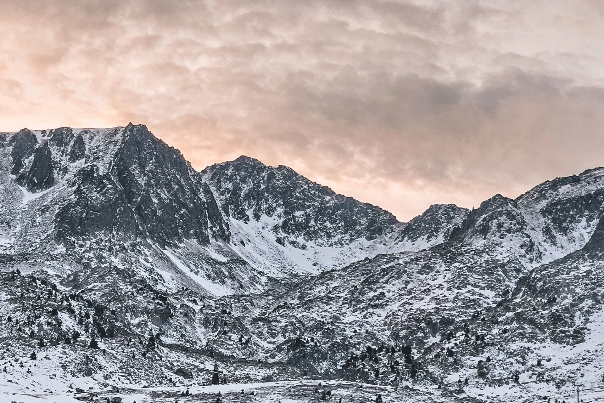 Snow-covered peaks in the Pyrenees Mountains in Andorra under a cloudy sky at sunset, with rugged alpine slopes and scattered trees, a landscape often explored during family travel in the high mountains of this small European principality.