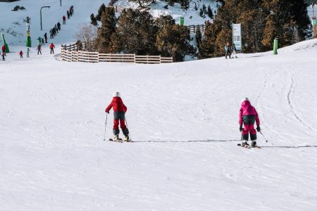 Two children skiing away from the camera down a wide beginner run, one in red and one in pink, with other skiers, a wooden fence and resort signage visible in the background.